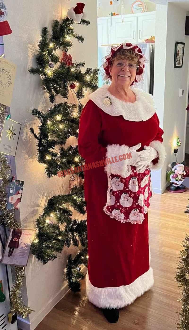Mrs. Claus for hire in Nashville standing beside a slim lit tree in a home hallway.