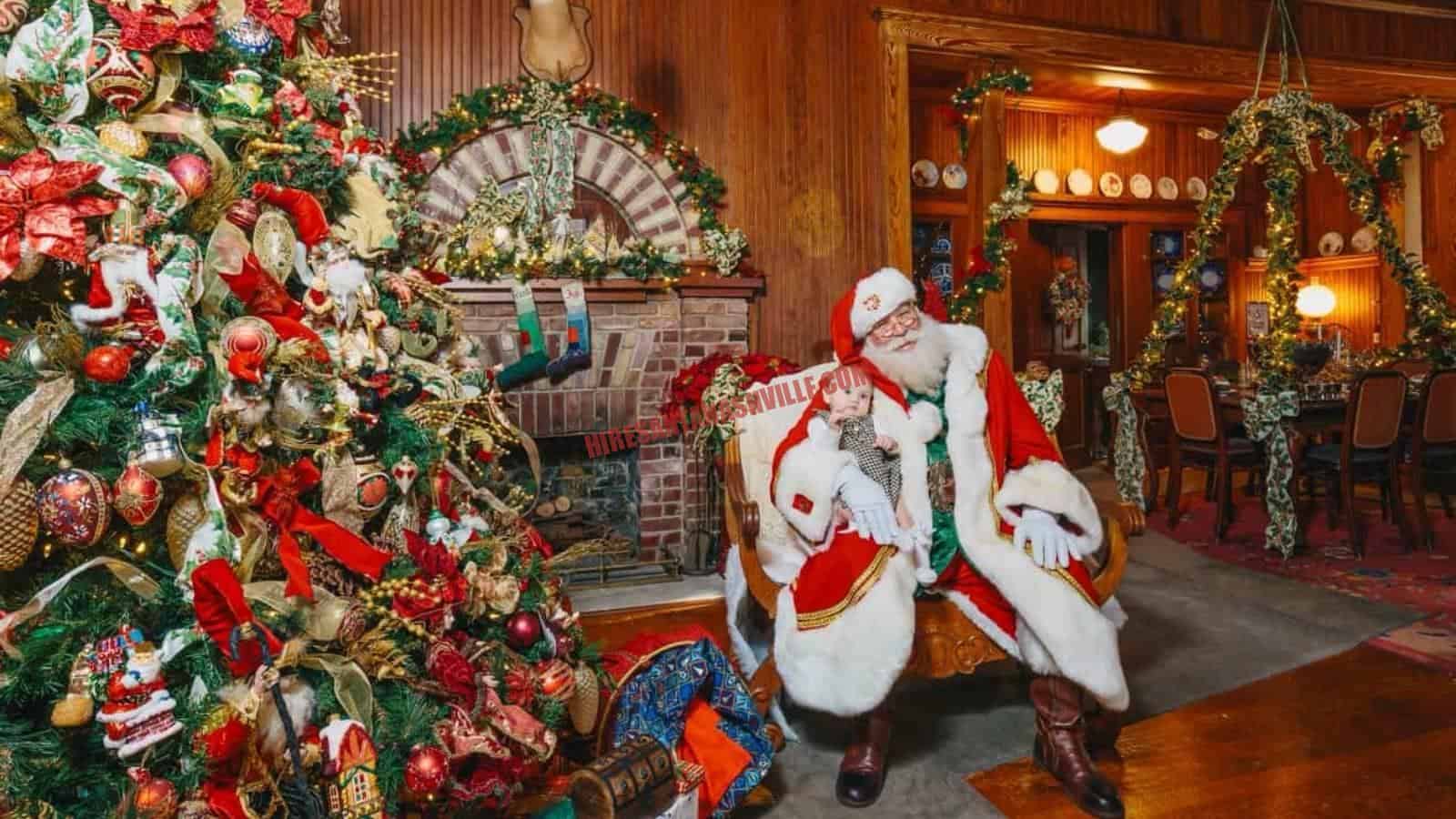 Santa Claus for hire in Nashville, TN seated by a fireplace and tree, holding a baby.